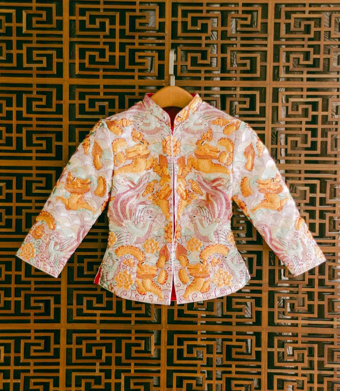 Hand-embroidered Chinese qun kwa wedding jacket on a hanger against a carved lattice screen, tailor-fitted and available to rent through Jenny Chiu Weddings